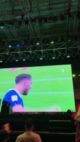 A wide screen displaying a soccer match is visible in the foreground. A man in a blue jersey is shown from behind, looking to his right. In the background, a large crowd of people are gathered outdoors at night, many of whom are holding up their phones and arms in excitement. The atmosphere appears to be festive, possibly at an outdoor event or concert.