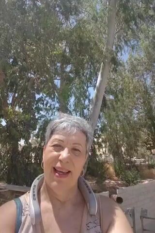 A smiling elderly woman with short grey hair and glasses is filmed in a medium close-up, facing the camera directly. She is wearing a light-colored sleeveless top and a white neck fan. She is outdoors on a sunny day, with several tall trees featuring green foliage and a clear blue sky visible in the background. The woman is actively speaking or singing throughout the short clip.