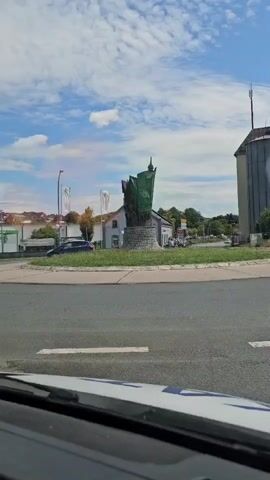 A car is driving around a roundabout. In the center of the roundabout is a sculpture that appears to be made of metal and stone. The sculpture is abstract and resembles a tower or a sail. It is green and brown in color. There are buildings and trees around the roundabout. The sky is blue with some clouds. In the foreground of the car's interior, a clear plastic container holds a few pieces of fruit, including apples and oranges.