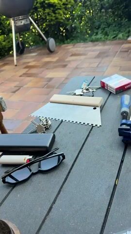 The video shows an outdoor scene on a tiled patio. There's a black kettle grill in the background, surrounded by green bushes. On a dark-colored wooden table, there are items like sunglasses, a lighter, a power bank, keys, and a metal tool with a wooden handle. Next to the table, there's a small wooden stool with a level, a hammer, and a can on top of it. At one point, a noise, like a revving car engine, is heard in the background. Then smoke starts rising from the grill.