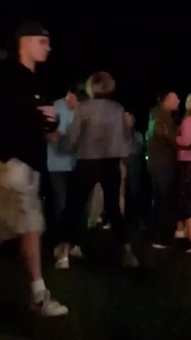 This video clip captures an outdoor nighttime event, possibly a party or festival, with people dancing. In the foreground, a man wearing a black hoodie and camouflage shorts walks from left to right, facing away from the camera. In the background, several couples are dancing. One couple in the center is dancing closely together, with the woman wearing a sparkly top and dark pants, and the man wearing a blue t-shirt and dark pants. To their right, another couple is dancing, with the man wearing a light-colored suit and a hat, and the woman wearing a pink dress. The overall scene is dimly lit with green stage lighting illuminating the area behind the dancers. The ground appears to be asphalt or a similar dark surface.