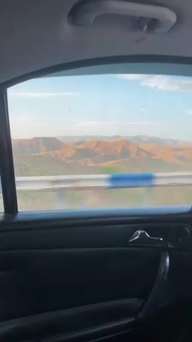 The video begins with a view through a car window. The interior of the car is visible in the foreground, showing a dark-colored door panel with a chrome door handle and a black fabric armrest. The window frame is also visible. Outside the window, a blue and white striped guardrail runs horizontally across the frame. Beyond the guardrail, a vast, arid landscape unfolds. Rolling hills, predominantly tan and orange in color, stretch towards the horizon. The sky above is a clear blue with some wispy clouds. The lighting suggests it is daytime, possibly late afternoon or early evening due to the warm tones of the landscape. No people or specific actions are discernable.