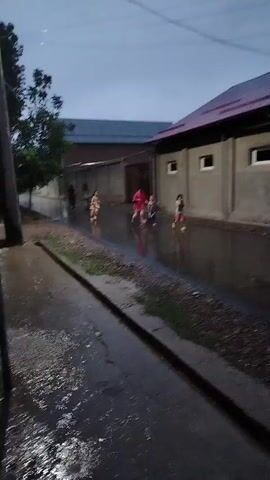 This is a video of children running on a wet street. It appears to be evening or overcast, as the lighting is dim. The street is covered in puddles, indicating it has recently rained or is currently raining. There are buildings on either side of the street, with a fence in the background. The children are running in various directions, some appearing to be playing. One child in the foreground is running towards the camera.