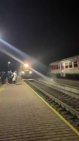 At night, a train arrives at a station. The train is white with red stripes and has a destination sign that reads "Kaunas VIA". There are passengers waiting on the platform. Another train, red and white, is parked on the adjacent track.