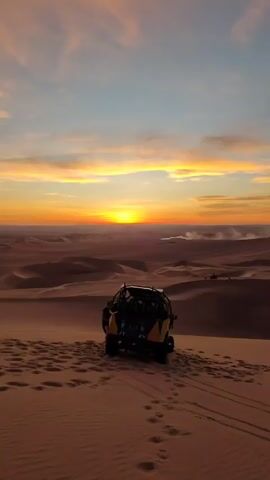 The video captures a desert scene at sunset. Initially, a dune buggy sits on a sand dune, with the sun setting over the desert landscape in the background. As the video progresses, the camera shifts to a red dune buggy.  Several people are visible on the dune, including a woman with her hair in a bun who is looking out at the landscape. Some people are sitting on the dune. Footprints are evident in the sand. The sky is colorful with hues of orange, pink, and blue.
