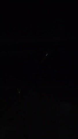 A drone flies across a dark, starless sky. The drone is a small, dark object with a bright green light on its underside, and what appears to be a red light or reflection along its body. The drone is in motion from the lower left corner of the frame to the upper right. In the lower left foreground of the frame, a thin, dark pole extends vertically, with a faint, wire-like object extending from its tip towards the drone. The environment is entirely dark, suggesting nighttime.