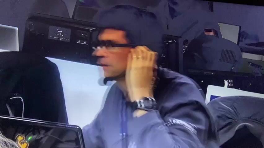 The video begins with a close-up of a man wearing glasses and a dark jacket, with his hand touching his ear. He is looking intently to the left, his expression one of concentration or concern. The background is dark and out of focus, suggesting an indoor setting with artificial lighting.

The scene then shifts to a wider view of a room filled with people seated at workstations, each equipped with multiple computer monitors. In the background, a large banner reads "SPACE CONTROL ROSWELL NEW MEXICO." The individuals appear to be engaged in monitoring operations, with some wearing headsets and others interacting with their screens. The overall atmosphere suggests a command center or a control room during a significant event.

The video then returns to a close-up of a person with short dark hair, hands clasped together in front of their face, with a concerned or anxious expression. A blue lanyard with white text is visible around their neck. In the background, another person is partially visible, also looking towards something out of frame. The lighting in this segment is dramatic, with highlights on the subject's face and a dark, shadowed background.