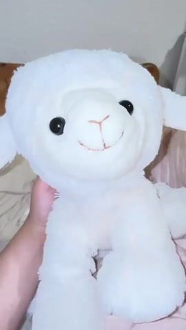 A close-up shot shows a white plush toy lamb being held by a hand. The lamb has black eyes and a smiling mouth with brown stitching. The background is blurred but appears to be a bed with a striped pink and white sheet. The video pans slightly to the right, revealing the lamb's ear.