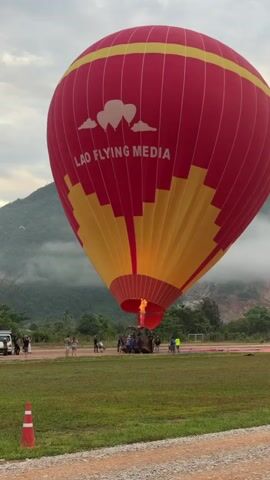 A red and yellow hot air balloon is being inflated on a grassy field with a mountainous, cloudy background. Several people are standing around the base of the balloon, and a vehicle is parked to the left. The balloon has the words "LAO FLYING MEDIA" printed on it in white lettering with cloud graphics. A flame is visible coming from the burner system, directed into the balloon's opening. A traffic cone is in the foreground. The time-stamped video shows the flame periodically intensifying as the balloon continues to inflate.