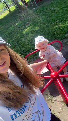 A young woman is holding a baby on a red merry-go-round in an outdoor park setting. The woman, with long brown hair, wears a white t-shirt with partially visible text including "morning" and a dark cap. She is smiling and looking at the camera, seemingly recording a selfie video. The baby, wearing a white hat and a light-colored outfit, is standing on the merry-go-round, holding onto a red bar, and smiling towards the camera with a joyful expression. The merry-go-round is slowly spinning. The background features green grass and out-of-focus trees under bright sunlight, indicating a clear, sunny day. Both the woman and the baby appear happy. The scene takes place in Красный Маяк, Russia.