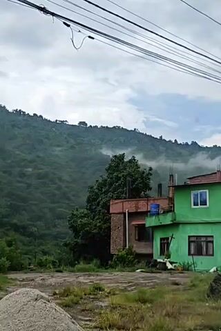 The video captures a street scene in what appears to be a mountainous region. In the foreground, a yellow dump truck is partially visible, loaded with what looks like gravel or construction material. The truck has text on its side, with "Lord Krishna" and a depiction of the deity visible. Further back, a vibrant green building stands out against a backdrop of lush green hills, partially obscured by mist or clouds. Adjacent to the green building is a brick structure with a red tiled roof. A small shop with a blue awning is situated in front of one of the buildings, displaying various goods for sale. A motorcycle covered with a black tarp is parked near the shop. Power lines crisscross the sky, and a street lamp is visible on a pole. The overall impression is one of a developing area with ongoing construction and local commerce set against a scenic, natural landscape.