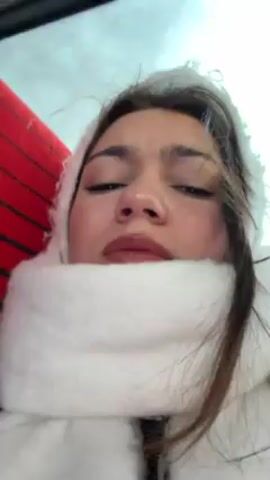 A woman is recording a video, likely of herself, as she appears to be bundled up for cold weather. She is wearing a thick, white scarf and a hood, possibly part of a jacket. Her eyes are partially closed, and she is moving her mouth as if talking, possibly singing along to music playing in the background. The environment seems to be indoors, maybe a vehicle, suggested by the presence of a red surface and a blue object in the background. The camera angle is low, looking up at the woman's face.
