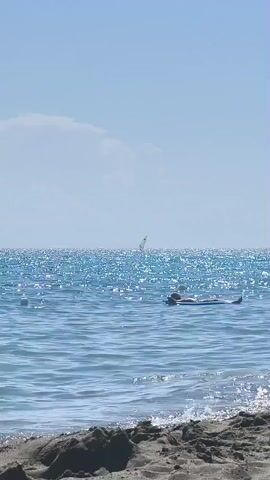 A person lies on an inflatable raft in the ocean. The water is blue and sparkles under a clear sky. In the distance, a sailboat is visible. Waves gently lap the shore in the foreground.