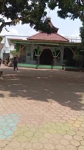 A person is walking from left to right across the screen. In the background, there is a mosque with a domed roof and an arched entrance. Trees with green leaves are visible in the foreground and on the sides of the mosque. The ground in front of the mosque is paved with hexagonal tiles, some of which are colored. The day appears to be sunny with shadows cast on the ground.