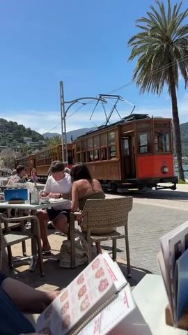 The video begins from a low angle, looking out from a restaurant towards the sea. In the foreground is a wicker table with a menu and a napkin dispenser. In the midground, a couple is sitting at a table, eating, with a red and brown tram passing behind them. In the background, there is a beach, sea, and mountains with buildings. The sky is blue with scattered clouds. The camera pans right, showing a man walking on the tiled walkway towards the beach. There are palm trees along the walkway. The beach has straw umbrellas and people relaxing.