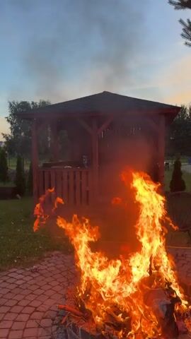 The video captures an outdoor scene at dusk in Dreiliņi, Latvia. In the foreground, a large bonfire rages with bright orange and yellow flames, casting a warm glow on the brick patio where it's situated. Behind the fire, a wooden gazebo with a dark roof and wooden railings is visible. Smoke from the bonfire drifts upwards into the twilight sky, which transitions from pale orange near the horizon to a deep blue.

The setting appears to be a backyard or park area. The ground is covered with green grass and the brick patio. In the background, there are trees and some houses suggesting a residential or semi-rural location. A few small, dark objects, possibly birds or debris, are scattered across the sky. The overall mood is serene, with the vibrant fire providing a focal point against the calm evening sky. There is no visible text in the video.