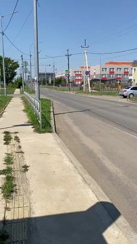 The video shows a view of a street from a sidewalk. On the right side of the road are several buildings with red roofs and colorful facades, likely residential buildings or a school. There are also cars parked along the road and on the side. A street light and utility poles with wires are visible. The sky is clear and blue with some scattered clouds. The scene appears to be a sunny day in a suburban or semi-urban area.