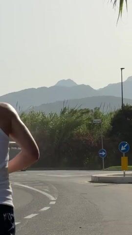 A young man wearing a white tank top and dark shorts is jogging on a paved road. He is facing away from the camera. In the background, there are lush green trees and bushes, with a hazy mountain range in the distance. The sky is bright and appears to be daytime. To the left, a dark colored SUV with a roof rack is parked. There are traffic signs visible, including a blue circular sign with a white arrow pointing to the right. The scene suggests an outdoor, possibly scenic, environment.