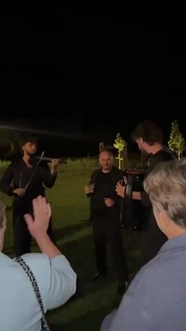 The video shows a group of people at an outdoor event at night. In the foreground, two women are visible from behind, with their backs to the camera. They appear to be clapping or gesturing with their hands. In the middle ground, three men are performing. One man on the left is playing the violin. Another man in the center is holding a drink and appears to be speaking or singing. The third man on the right is playing the accordion. The men are all dressed in dark clothing. The background is dark, with some trees and lights visible. The ground is covered in grass.