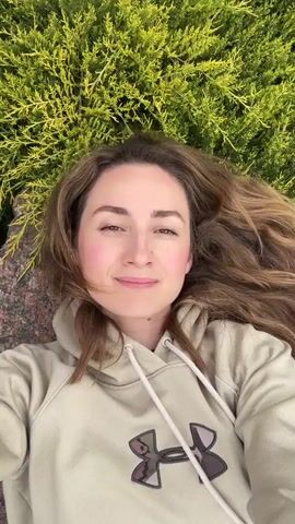 A woman lies on her back, smiling up at the camera. She wears a tan hoodie with a dark camouflage design. Her brown hair is spread out around her head. She is positioned between a green, densely-leafed bush and a large, rough-hewn red rock formation. As the camera tilts up, more of the rock formation is revealed, with more of the green foliage surrounding it.