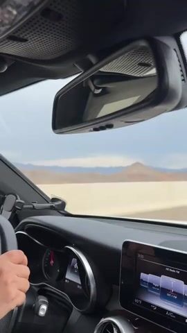 The video shows the interior of a car, with the dashboard and rearview mirror prominently featured. Through the windshield and reflected in the mirror, a desert landscape unfolds. In the foreground, a hand is visible on the steering wheel. The dashboard displays a digital clock showing "3:33" and what appears to be a radio interface with "HD Radio FM" and station information including "Vegas 106.9 FM" and "107.1 FM." The weather appears overcast, with a cloudy sky and distant mountains. The overall mood suggests a road trip or commute through a dry, arid region, possibly during the daytime. The visible text provides context about the radio stations being tuned, indicating a location near Las Vegas, Nevada. The scene is static, with no discernible movement within the vehicle itself, suggesting the car is in motion.
