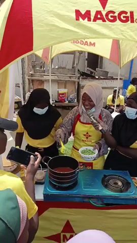 A young woman wearing a hijab and a yellow apron with a Maggi logo is cooking over a portable stove. She is stirring a pot with green peas. To her left and right are two other young women also wearing hijabs and yellow shirts with the Maggi logo. They are wearing face masks. A yellow and red tent with the Maggi logo is overhead. There are other people in the background, some wearing yellow shirts. A person in the foreground is holding up a phone to record the cooking demonstration. The event appears to be an outdoor promotion for Maggi products.