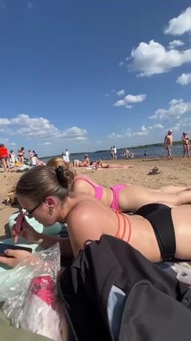 A sunny day at the beach with several people sunbathing on the sand. In the foreground, two women are lying on their stomachs on beach towels, facing away from the camera. The woman closer to the camera is wearing black bikini bottoms and an orange bikini top, and she is looking at her phone. The woman behind her is wearing pink bikini bottoms and a pink bikini top. In the background, other people are visible on the beach and in the water, some standing and some lying down. The sky is blue with scattered white clouds, and there is a body of water in the distance. There is a can of soda on the beach towel in the foreground.