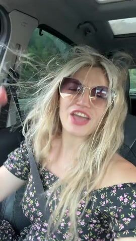 A woman with long blonde hair and sunglasses is sitting in the passenger seat of a car, with her hair blowing in the wind. She is wearing a floral off-the-shoulder dress and a seatbelt. The car appears to be moving, as indicated by the motion blur and the wind in her hair. The background is out of focus but suggests an outdoor environment with trees. The video is shot from a selfie perspective, with the woman looking directly at the camera.