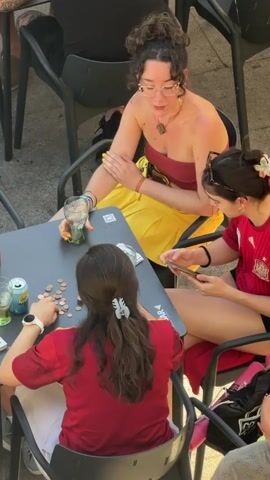 This media file shows a group of young women sitting at a table outside a cafe. The scene is bright and appears to be daytime, likely sunny given the shadows. The women are casually dressed, with two of them wearing red shirts. One woman in the foreground has her back to the camera, with her hair in a bun and a white smartwatch on her left wrist. She is reaching for coins scattered on the table.

In the center, a woman with curly dark hair tied up and wearing glasses, a red bandeau top, and a bright yellow skirt is gesturing with her left hand. She is also wearing a necklace and several bracelets. To her right, another woman with dark hair in a bun, adorned with a flower, is looking at her phone. She is wearing a red shirt with the Spanish flag crest visible on the sleeve.

The table is dark and set with drinks, cans, and a significant amount of small copper coins. The arrangement of the coins suggests they might be engaged in a game or calculation. The setting appears to be an outdoor seating area of a cafe or restaurant, with dark plastic chairs and a paved ground visible. The presence of Spanish national colors on the clothing and the mention of Las Rozas de Madrid, Spain, provide strong locational context. The overall mood seems relaxed and social, typical of people enjoying a casual outing.