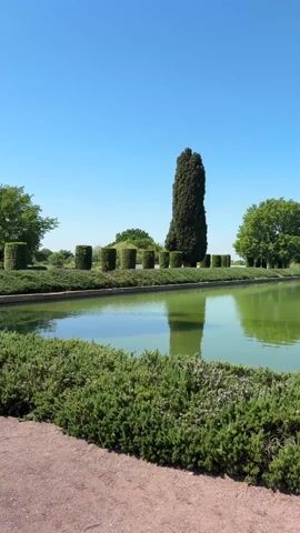 The video pans across a manicured garden in Rome, Italy. The camera begins facing a long, rectangular pool surrounded by trimmed hedges and trees in cylindrical shapes. The camera then pans right to reveal a wider walkway. A group of four people are standing in the walkway and a woman with long red hair is kneeling on the side taking photos. In the background is a large grass field with trees scattered throughout. A large, ancient building is visible in the distance. The sky is clear and blue, with a few clouds.