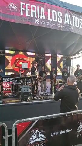 A young male singer in a black hoodie performs on a stage at “Feria Los Altos.”  He stands behind a microphone, with a keyboard and a laptop on a stand to his left.  Behind him, several band members stand ready, one with a saxophone and another with a trombone.  Behind the band, a large LED screen displays shifting colors and graphics.  In front of the stage, a man holds up a phone and records the performance. A red and black banner with the “Feria Los Altos” logo is visible in the foreground. The scene appears to be outdoors, possibly at a festival or event.