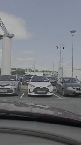 A grey SUV and a white hatchback are parked in a lot. A man in a pink shirt and jeans walks toward the white hatchback. A woman in a blue tank top and jeans follows him. They walk past the front of the white hatchback and out of frame.