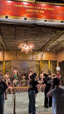 The video shows a group of people watching a performance on a stage in what appears to be a cultural center.  The stage is decorated with bamboo and has a sign reading "TEH DAR" in the center.  On the stage, shirtless male performers are playing drums and other instruments.  The audience members are standing and watching the performance, some are taking photos.  The space is lit by a large, decorative light fixture hanging from the bamboo ceiling.  Above the stage is a sign that reads “Trung tam bieu dien nghe thuat, Lune Hoi An”.