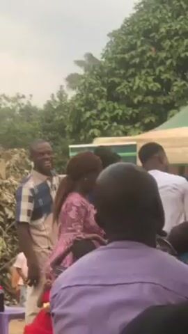 The video shows an outdoor gathering of people, with a focus on a group near a tent. The tent has green and white stripes on its top. Several adults are interacting with children in the group. One man, wearing a blue and white checkered shirt, is smiling and appears to be engaged with the children. Another man wears a white long sleeve shirt. Some of the children are dressed in colorful attire, while others are in casual clothing. Behind the group is a banner with text that reads "Diana Women of Anna." The environment is green and leafy, with trees visible in the background. The camera view is partially obstructed by the backs of seated people, mainly dressed in dark colors, presumably members of the audience or onlookers.