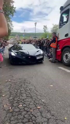 Here is a description of the video:
The video shows a dark-colored Bugatti Mistral convertible parked on a cobblestone street. There is a large crowd of people lining both sides of the street and a semi-truck parked directly in front of the Bugatti. An individual in an orange safety vest is standing by the front right tire of the truck. Buildings can be seen in the background, along with some trees. The sky is visible and it's a partly cloudy day. The car has a license plate that begins with "W".