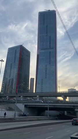 The video captures a ground-level view of urban architecture featuring a multi-level highway interchange in front of two prominent skyscrapers. The skyscrapers are predominantly glass with vertical red accents and are positioned against a backdrop of a partly cloudy sky with a hint of sunset. A white and yellow taxi with visible license plate is moving on the road in the foreground. Pedestrians can be seen walking along the sidewalk beneath the highway. The video maintains a static perspective with no camera movement.