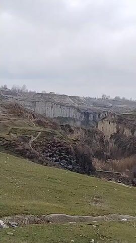 The video shows a landscape on an overcast day. It pans across a green, grassy field towards a steep, eroded canyon with bare trees. The camera then transitions to a muddy, uneven dirt road with scattered patches of grass and standing water. Concrete poles, some supporting a rudimentary fence, line the side of the road. The sky remains consistently gray and overcast throughout the video. The scene lacks any human presence or activity.