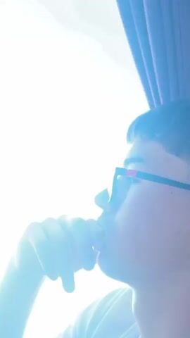 The video features a young man in profile, seated inside a vehicle, looking out a window on a bright, sunny day. He wears a white t-shirt and black sunglasses with red accents, with his right hand resting near his mouth, suggesting contemplation or observation. Initially, the exterior view is overexposed and blurry, but it quickly clarifies to reveal a rugged, rocky hillside under a clear blue sky. The rocks are light-colored and appear stratified, with sparse green vegetation in places. The vehicle is moving, indicated by the passing scenery. A brown curtain is visible on the right side of the window. The scene depicts a calm journey through a sun-drenched, mountainous or hilly landscape. A female voice speaking in a foreign language (likely Slavic) can be heard in the background, mentioning "Orthodox Christians" and "Muslims."