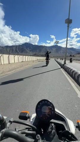 The video shows a person standing on a moving motorcycle, with their arms outstretched. The motorcycle is on a paved road in a mountainous area with a clear blue sky and white clouds. The camera is mounted on another motorcycle, following behind the first one. The person on the first motorcycle is wearing a black jacket and helmet. The road is bordered by a low concrete wall and a sandy area with sparse vegetation. In the background, there are large mountains. There are other motorcycles in the distance, also on the road. A solar panel is visible on a pole near the road.