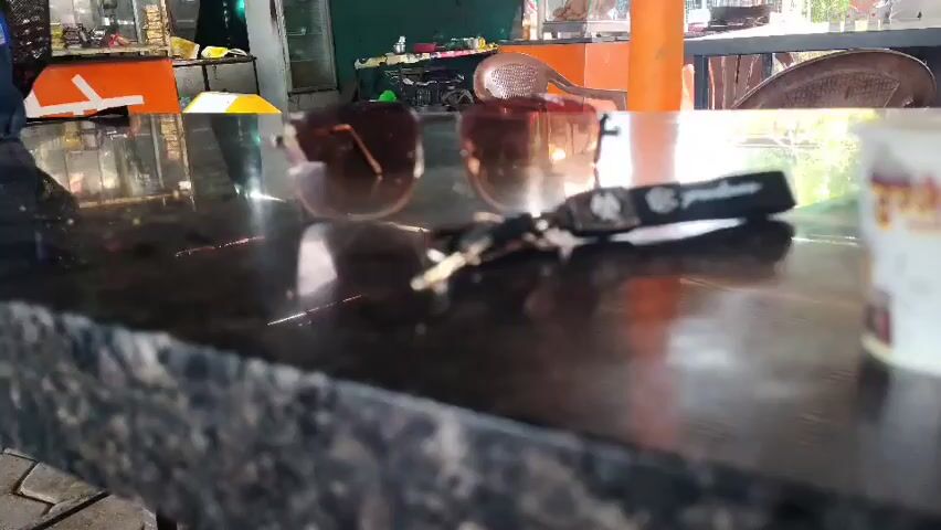 The video shows a close-up shot of a dark granite countertop. On the countertop are a pair of sunglasses, a set of keys with a black keychain, and a small paper cup. The cup has a red heart and the words "I My Ujjain" printed on it, along with some text in what appears to be Hindi script above. The background is blurred, but appears to be an outdoor or semi-outdoor setting with tables, chairs, and some foliage. A person is briefly visible in the background, wearing a blue shirt and dark pants, walking away from the camera. The lighting suggests it is daytime.