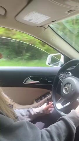 A person is driving a silver Mercedes Benz on a winding road through a forest. The person is wearing a grey hoodie and has manicured nails. The car's dashboard has a touchscreen display with various apps. Lush green trees line both sides of the road.
