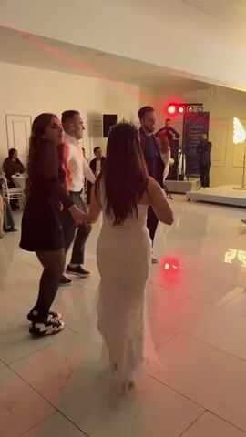 In a brightly lit reception hall, several people are dancing on a shiny, white tiled floor illuminated with vibrant purple, blue, red and green lights. The dancers include a woman in a white gown and tiara holding hands with a woman in a short black dress and leggings. Two men stand beside them, also holding hands. A band is set up in the background on a raised platform, with several musicians and sound equipment. To the right of the band, a sign is visible, along with a lit decorative butterfly. People are seated at tables in the background, and a man with a camera is actively recording the dancers. The walls are white and the room has a festive, celebratory ambiance.