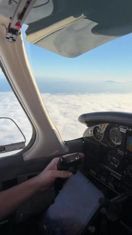 The video begins with a first-person view from inside an airplane cockpit. A hand is visible holding a radio or control device. Outside the cockpit window, a vast expanse of white clouds stretches to the horizon under a clear blue sky. In the distance, faint mountain ranges are visible through a hazy atmosphere. The view remains focused on the clouds and the distant mountains for the duration of the clip, offering a serene aerial perspective.