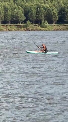 A single person is paddling a light-colored stand-up paddleboard on a body of water, likely a river or lake. The person is kneeling on the board, using a paddle to propel themselves forward. They are wearing a black top. The water is choppy, and splashes are visible around the paddle as it enters the water. In the background, a dense forest of green trees lines the shoreline. The sky is overcast.