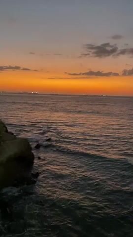 The video captures a serene sunset over the ocean. The sky is a gradient of deep blue at the top, transitioning to a vibrant orange band just above the horizon, with streaks of pink and purple. Scattered clouds add texture to the sky. The ocean below reflects the colors of the sunset, with waves gently lapping at a rocky shoreline on the left side of the frame. Several distant lights, possibly from ships or shorelines, are visible on the horizon. The overall atmosphere is peaceful and tranquil.