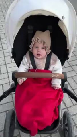 The video shows a baby in a stroller. The baby is wearing a beige hat and a patterned sweater, and is wrapped in a red blanket. The stroller has a white canopy and black frame. The baby looks around and at the camera. The stroller is on a paved surface with a repeating brick pattern.