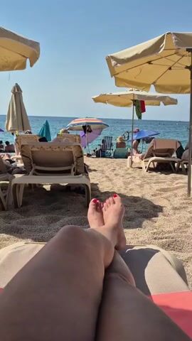 The video opens with a low-angle first-person perspective of a person lying on a beach chair. Their legs are crossed at the knees, and their feet are visible with red painted toenails. The beach is made of small pebbles and sand. In the background, several beach umbrellas and lounge chairs are set up. Some people are visible in the distance, relaxing on the beach and near the water. The ocean is calm and blue under a clear sky.