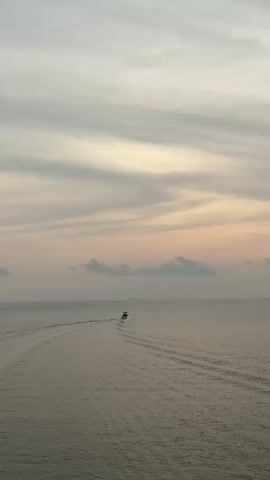 The video opens with a distant view of a small boat moving across a body of water, leaving a wake behind it. The sky is overcast with a hint of pink in the clouds, suggesting either sunrise or sunset. As the camera pans right, it reveals a rocky breakwater with a small structure, possibly a navigational aid. The sun, still hidden behind the clouds, casts a bright glare on the water. In the distance, there appears to be a larger ship or island. The scene remains still, capturing the quiet vastness of the sea under the cloudy sky.