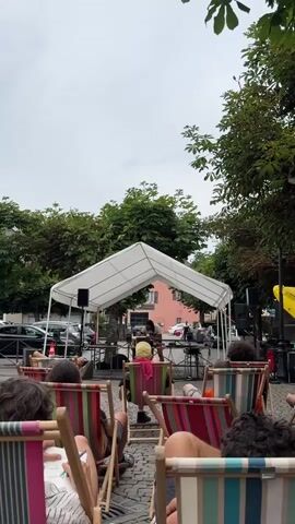 The video shows an outdoor event with a performance under a white canopy. Several people are seated in brightly colored lounge chairs, facing the canopy. A person is visible behind a table with electronic equipment under the canopy, seemingly performing. Trees line the background, and a building is visible behind the canopy. Cars and streetlights are also present in the background, suggesting an urban or town setting. The overall atmosphere appears relaxed and casual, possibly a public gathering or a small concert.