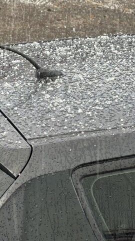 The video shows the roof of a gray car covered in small hailstones. Rain is falling in sheets, blurring the background, which appears to be a concrete or paved surface. The hailstones are densely packed on the car's roof, and water is running off the car's surface. The car has an antenna and appears to be parked.